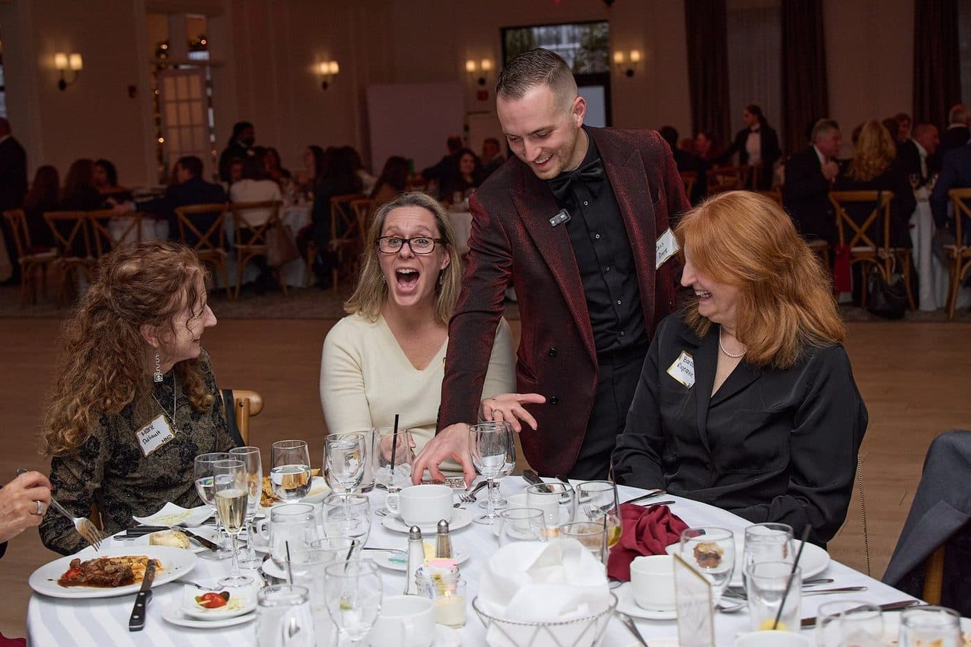 A group of women and a man at a banquet table, sharing a laugh with more guests in the background.