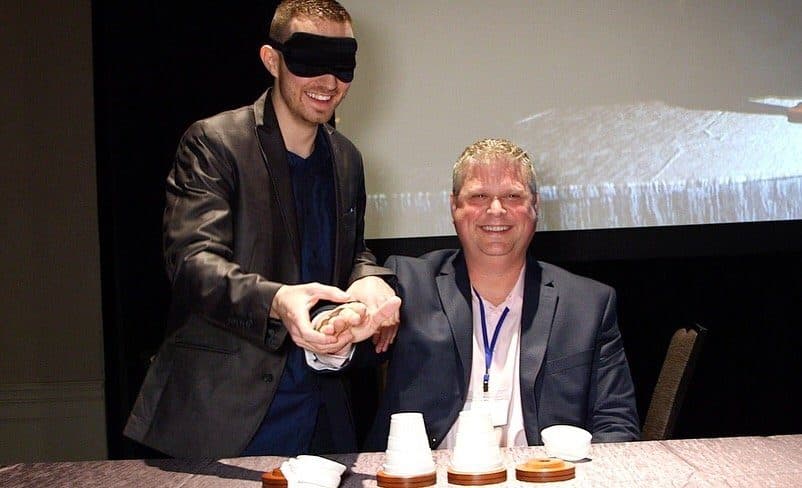 A man wearing a blindfold performs a coin trick for a smiling man sitting at a table during a signing event.