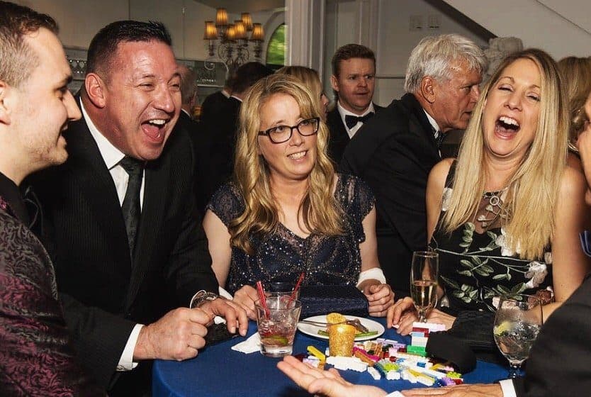 Group of five adults in formal attire enjoying conversation and drinks at a social event in a dimly lit bar or lounge.