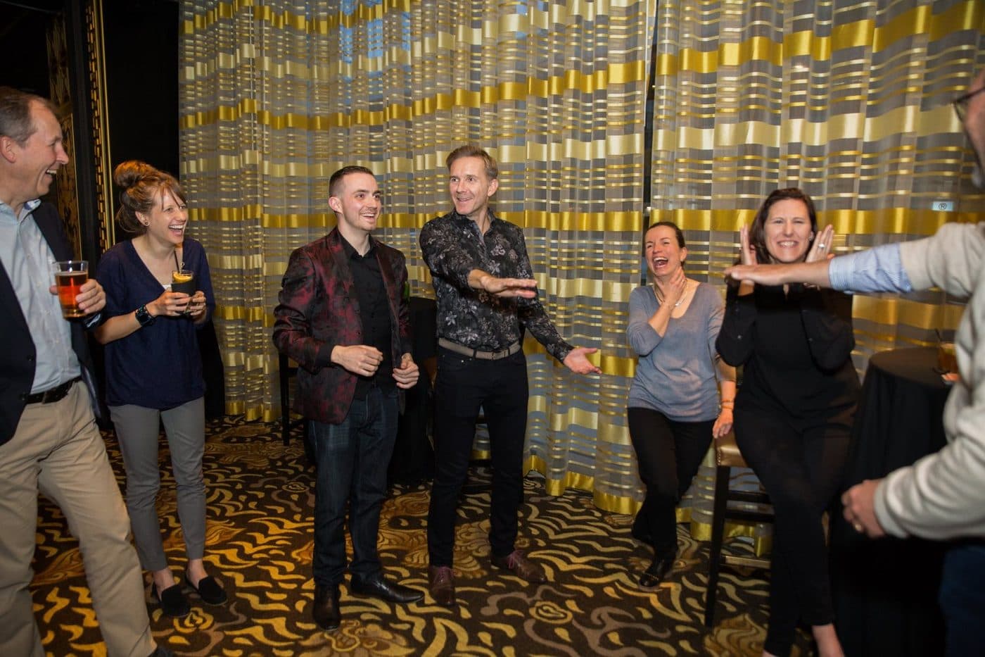 Group of people laughing and socializing at a party in front of gold decorative panels, some holding drinks.