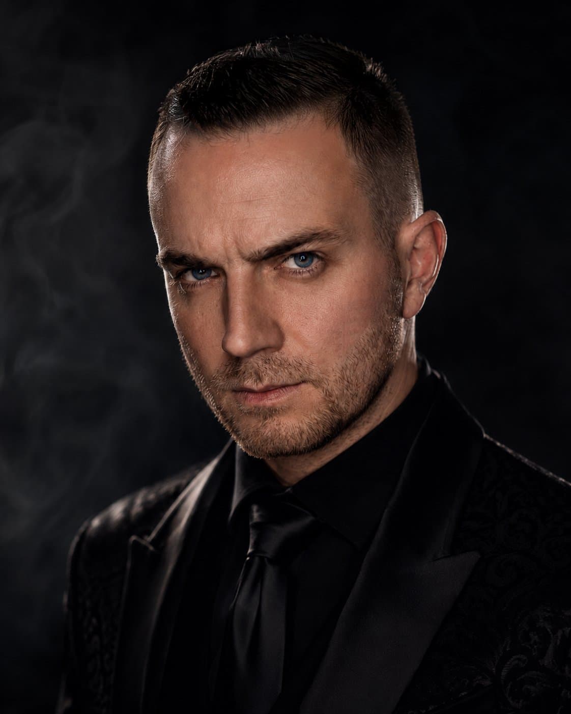 Chris Dare, NYC corporate magician and mentalist, in a black jacket and tie against a dark smoky background