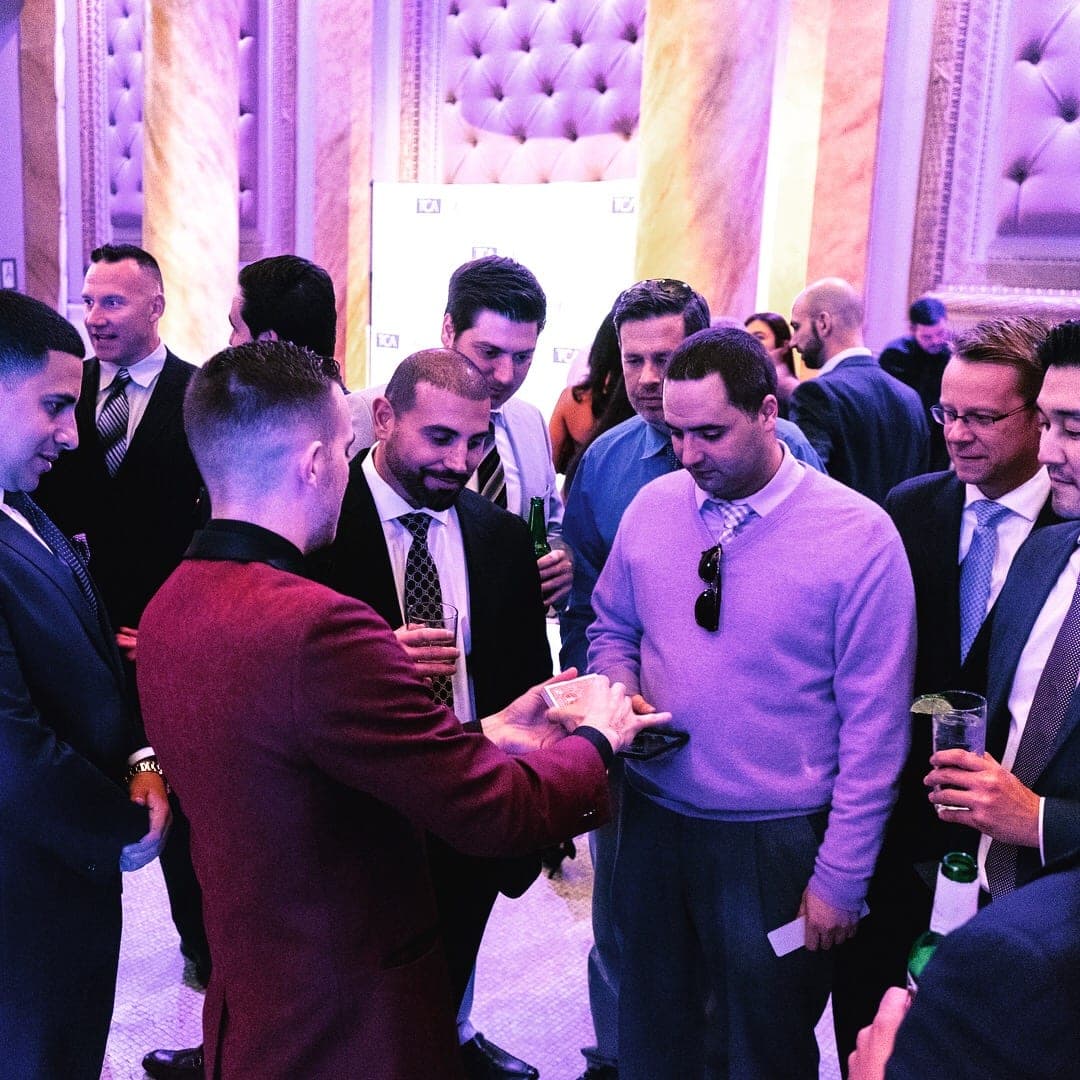 Businessmen crowding around Chris Dare as he reveals a chosen card during a corporate cocktail hour
