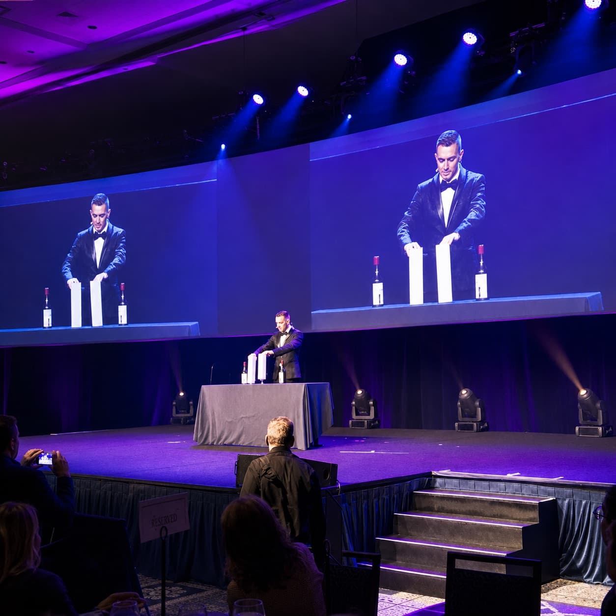 Chris Dare performing a stage magic and mentalism show for a corporate ballroom audience, shown on two large screens